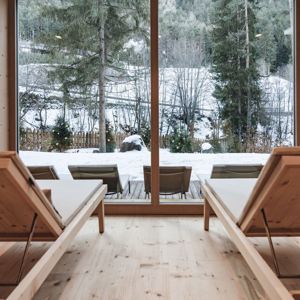 Sensoria Dolomites. Kraftort. Winterzauber. Blick aus einem Raum mit Holzliegen auf verschneite Landschaft und Tannenbäume