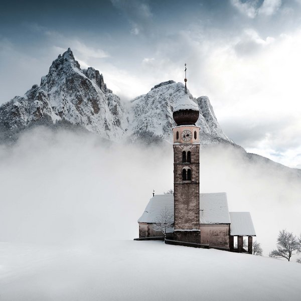 Sensoria Dolomites. Kraftort. Winterzauber. Kirche im verschneiten Tal vor nebelverhangenen Bergen