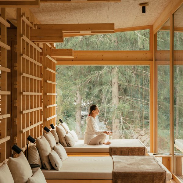 Sensoria Dolomites – Luxury for the Soul Woman relaxing on lounge bed in wooden room with large window and forest view
