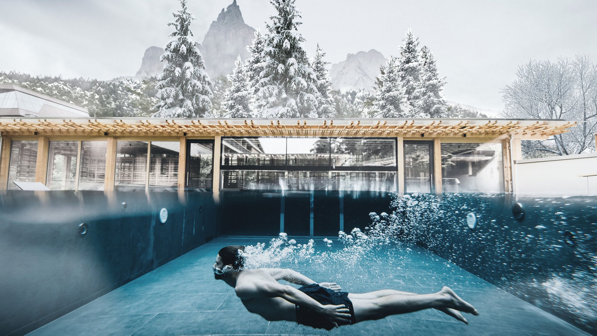 Sensoria Dolomites. Kraftort. Winterzauber. Mann schwimmt unter Wasser in Pool mit Blick auf verschneite Berge und Bäume