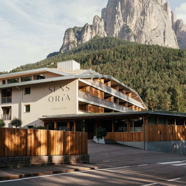 Sensoria Dolomites – Luxury for the Soul SENSORIA hotel with wooden facade in front of forested mountain in the Dolomites