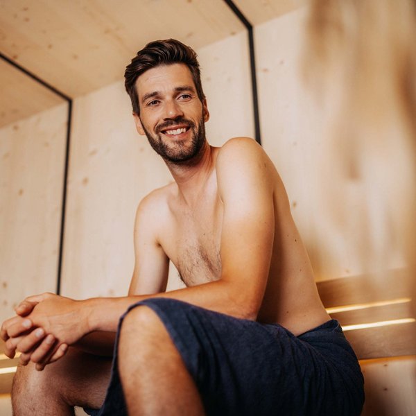 Sensoria Dolomites. Kraftort. Winterzauber. Lächelnder Mann sitzt in Handtuch in Sauna aus Holz