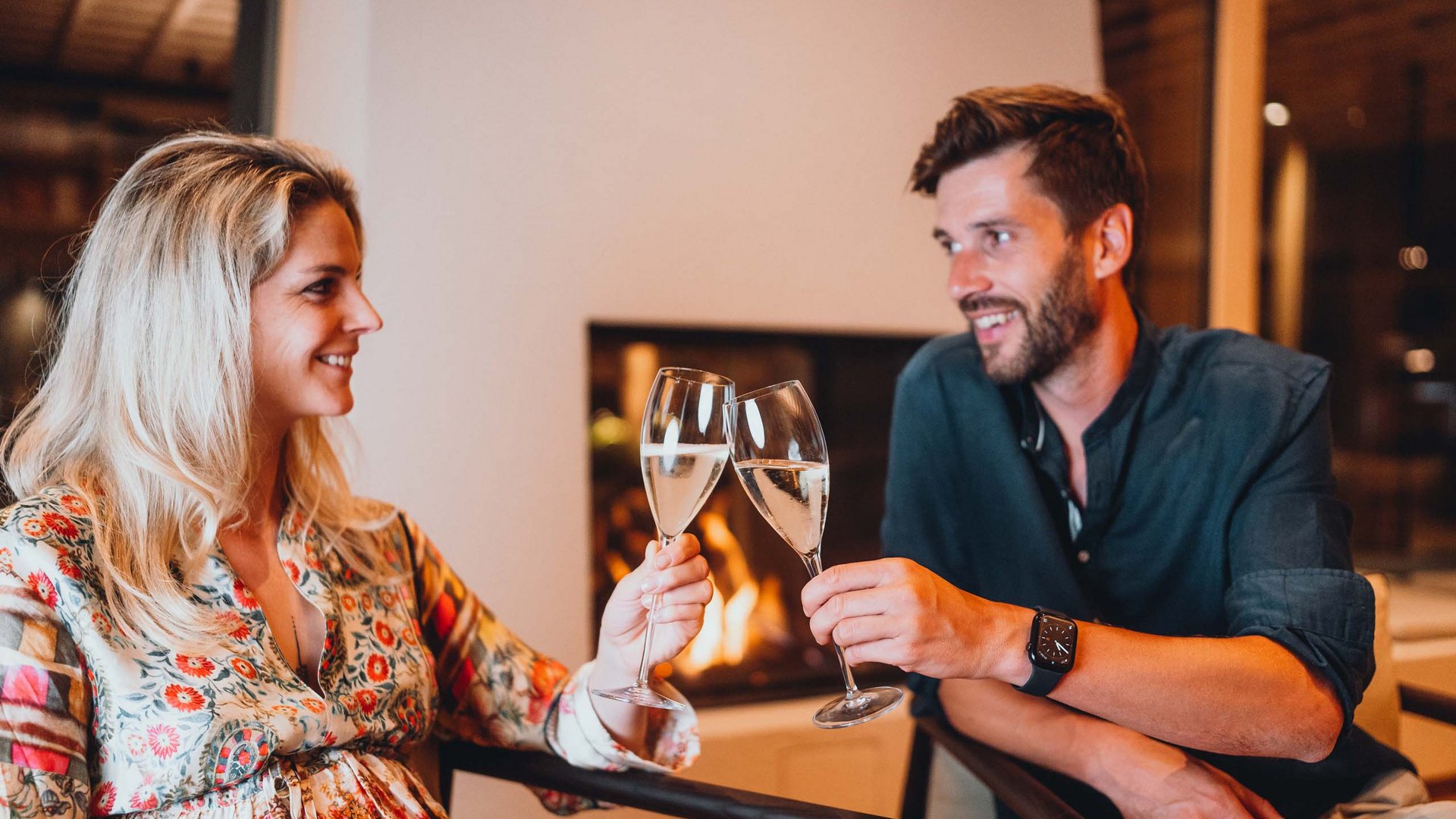 Sensoria Dolomites. Kraftort. Winterzauber. Paar stößt mit Sektgläsern vor gemütlichem Kamin an