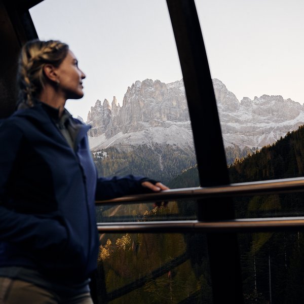 Sensoria Dolomites – Luxury for the Soul Frau blickt aus Fenster auf bewaldete Berge und Felsgipfel bei Sonnenuntergang