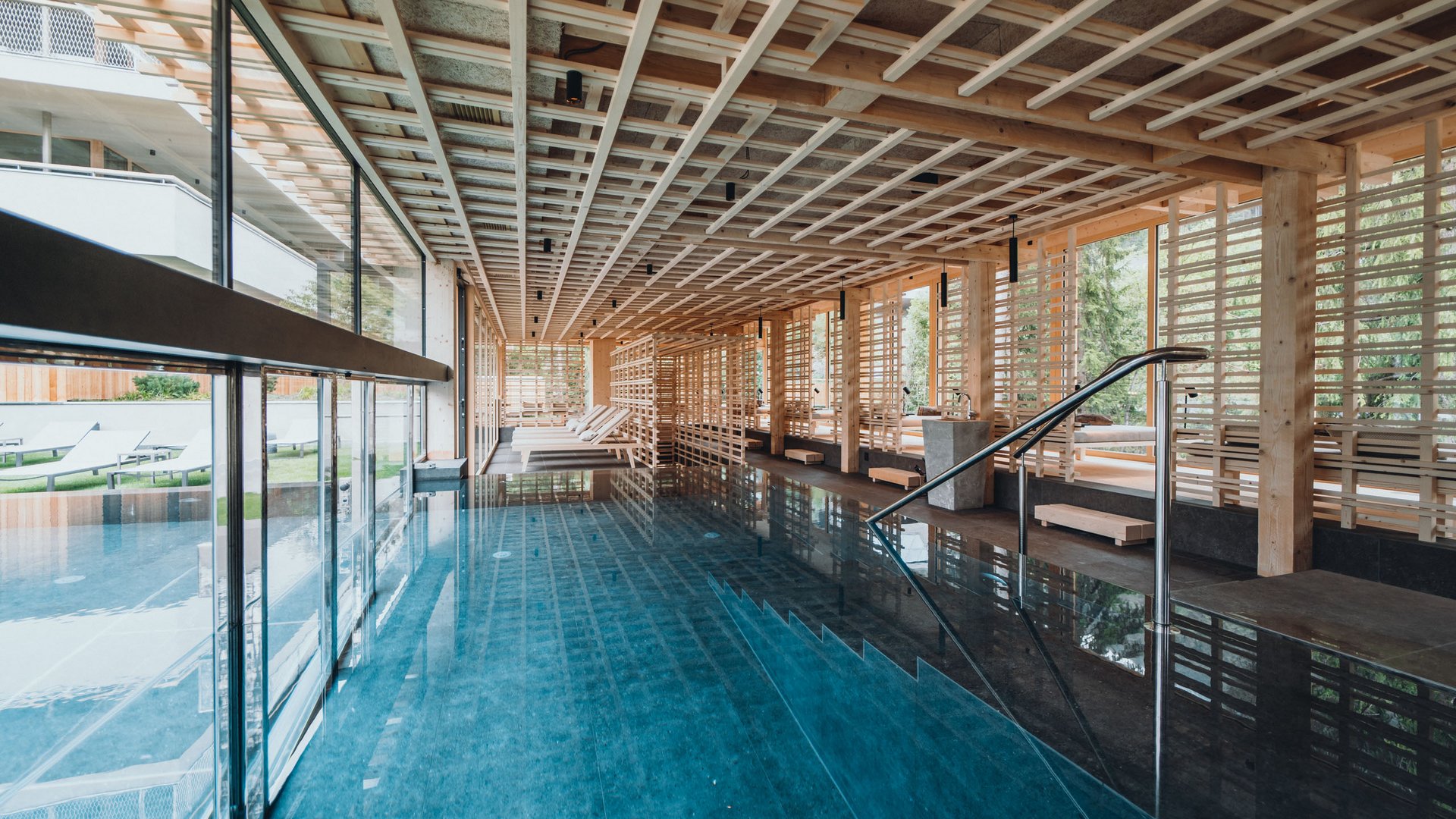 Sensoria Dolomites – Luxury for the Soul Modern indoor pool with wooden elements and lounge chairs by large glass windows