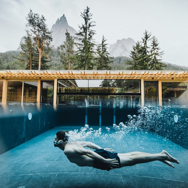 Sensoria Dolomites – Luxury for the Soul Man swimming underwater in pool in front of modern house with mountain backdrop