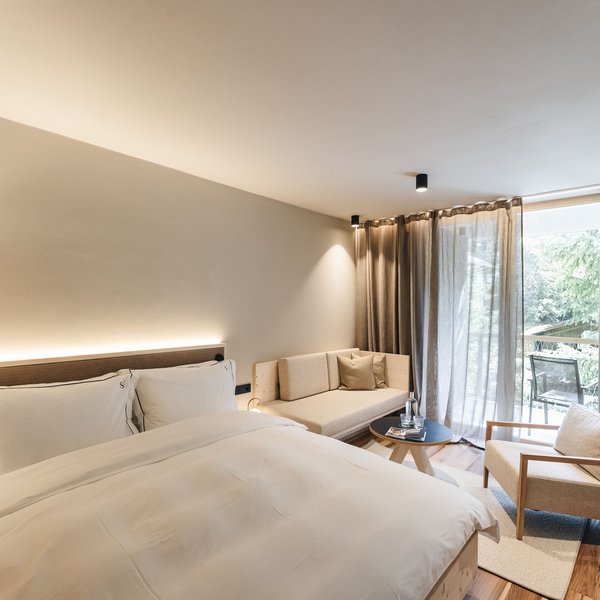 Sensoria Dolomites. Kraftort. Winterzauber. Helles Hotelzimmer mit großem Bett, Sofa, Sessel und Balkon mit Bäumen